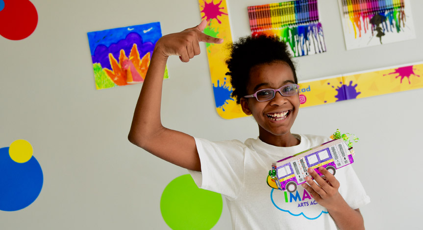 A girl with glasses is smiling while pointing at her paper bus she made during a Crayola imagine arts academy class